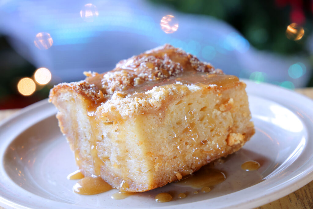 Close up of bread pudding straight off the buffet. There are some lights in the background and rum sauce drizzling down the piece.